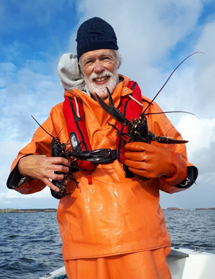 Foto på fiskare med orangefärgad galonjacka stående i en öppen båt på havet och som visar upp två humrar